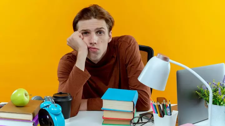 Сумний хлопець сидить за столом із книжками та ноутбуком